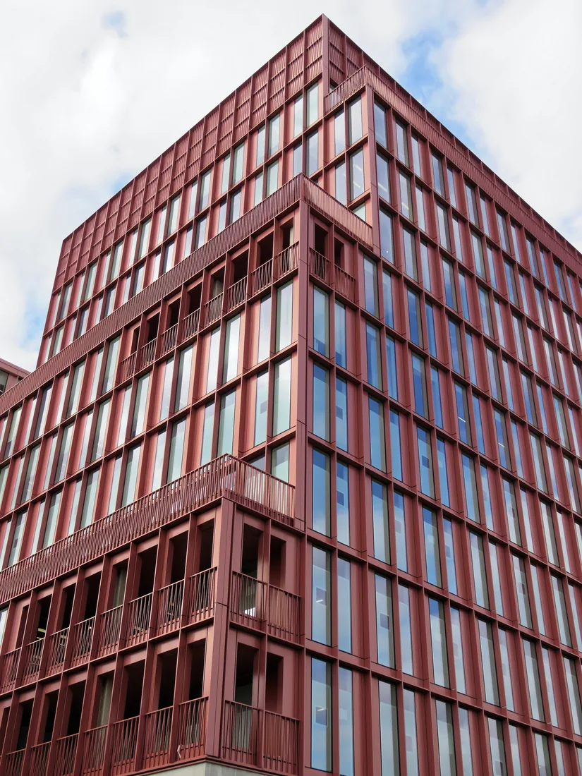 A tall red-framed office block with rows of tall windows and balconies, seen from below against a cloudy sky.