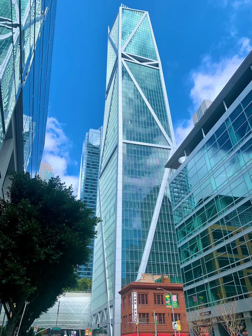 A slender glass skyscraper with white diagonal bracing, seen from street level above a small red-brick building.