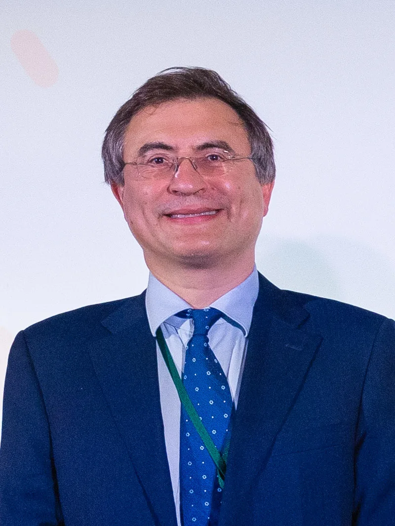 Chris Hohn, in glasses, a navy suit and a blue spotted tie, smiling against a pale background.