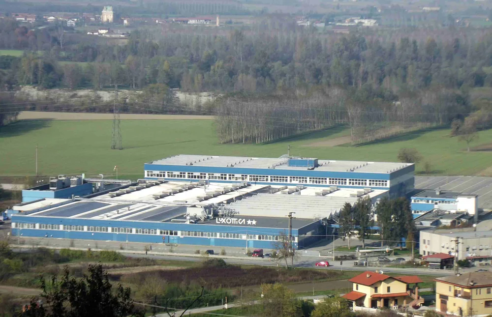A large blue-and-white factory building with the Luxottica name on its roof, set among fields and trees.