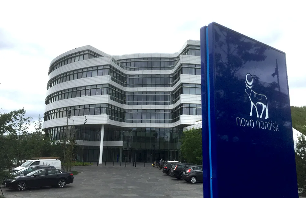 A curved white office building with a tall blue Novo Nordisk sign in the foreground.