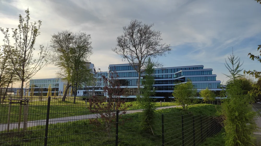 Modern glass-fronted office buildings of the Aldi Nord campus behind a fence and young trees.