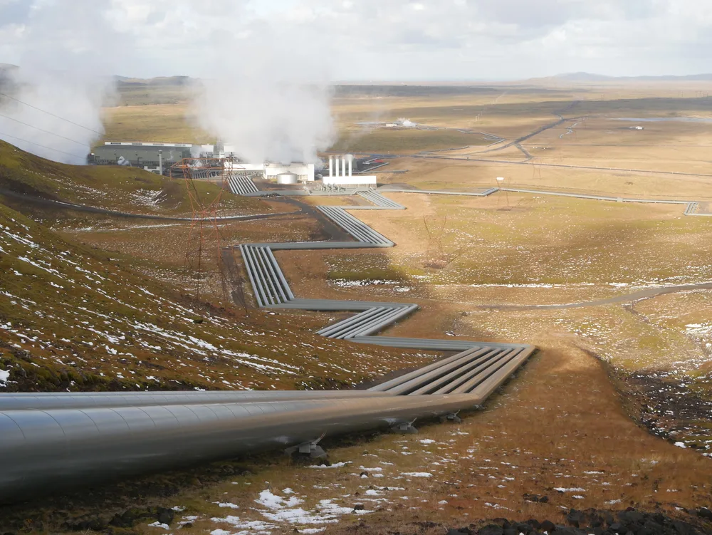 Silver steam pipelines zigzag across a moorland valley towards a geothermal power plant releasing white steam.