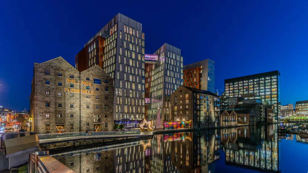 Lit office blocks and an old stone mill building reflected in the water of a dock at night.