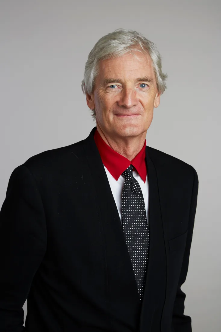 James Dyson, white-haired, in a black jacket, red-collared shirt and spotted tie, against a grey background.