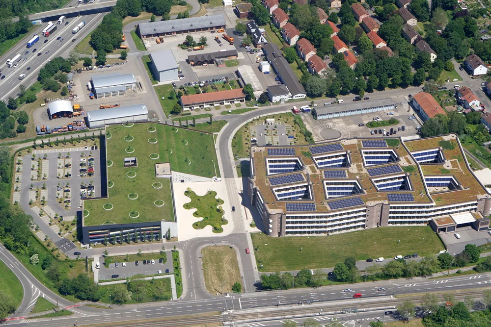 Aerial view of a large, low office complex with solar panels and green roofs beside a main road in Karlsruhe.