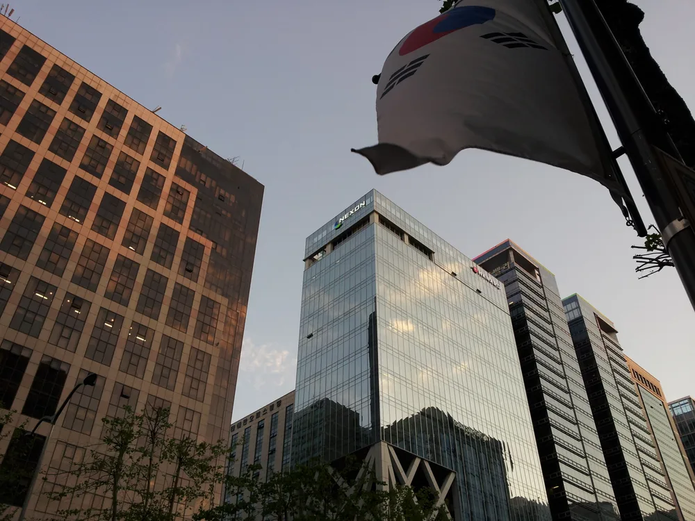 A glass office tower with the Nexon logo near its top, seen from street level beside a South Korean flag.