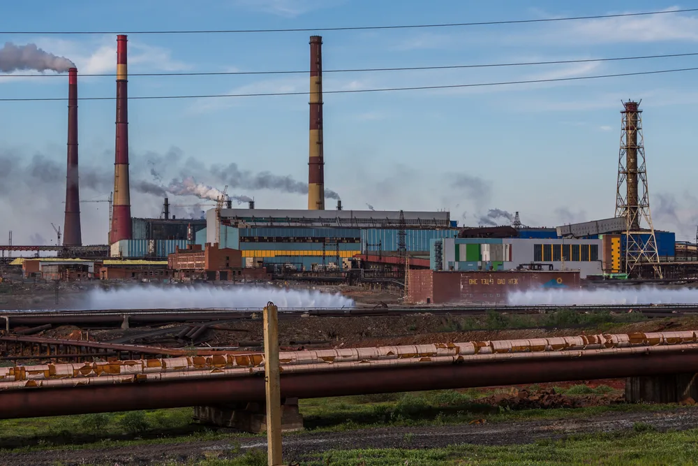 The Nadezhda smelter in Norilsk, with tall striped chimneys, steaming pipes and industrial buildings under a blue sky.