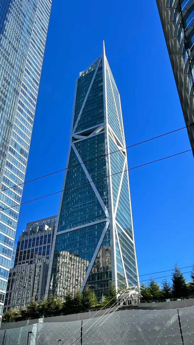 A tall glass skyscraper with white diagonal bracing rises against a clear blue sky between other office buildings.