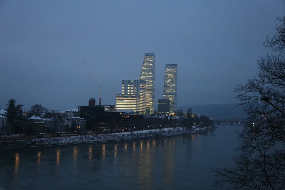 Two lit office towers rise behind snow-covered houses on the bank of a wide river at dusk.