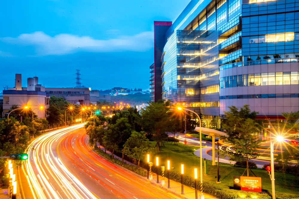 A glass-fronted TSMC factory building at dusk beside a curving road streaked with car light trails.